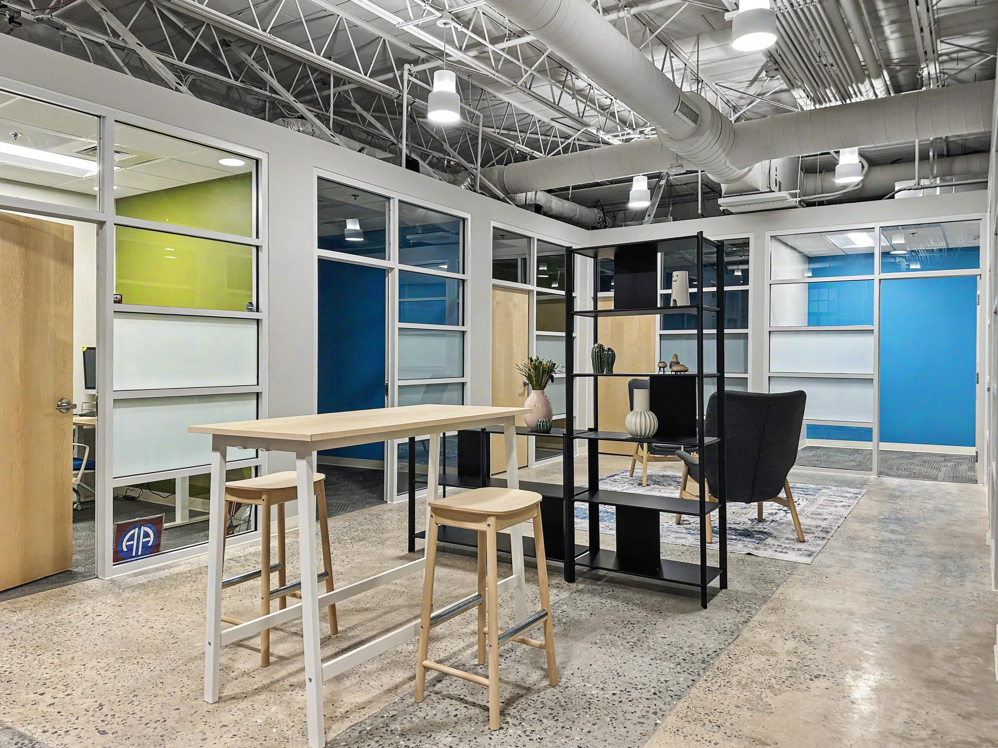 Private offices with glass panel walls, with a common area featuring a standing high-top table with stools for quick chats an Private offices with glass panel walls, with a common area featuring a standing high-top table with stools for quick chats an